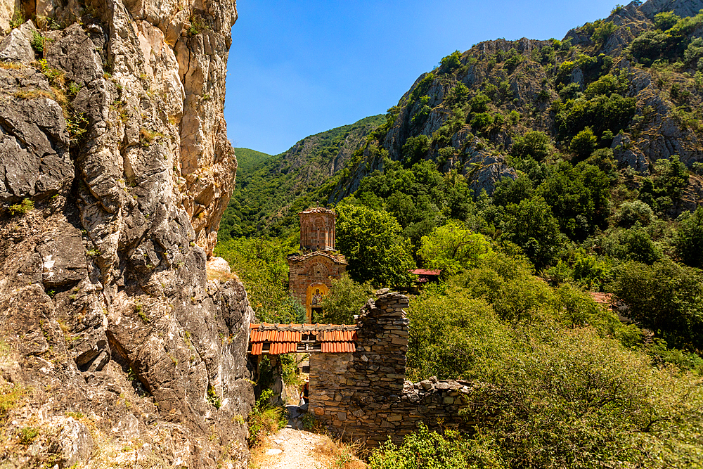 Horizontal eye-level shot showing a stone pathway along a cliffside leading to the remains of an old church with a red-tiled roof, surrounded by dense greenery and mountains at Matka Canyon.