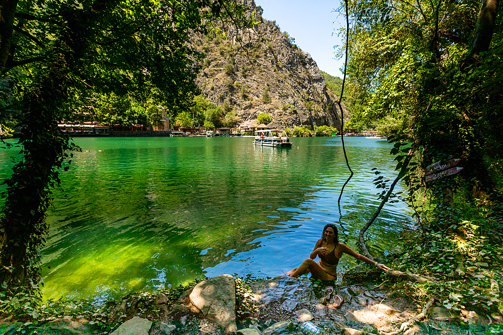 Horizontal eye-level shot framed by overhanging trees, showing a woman sitting at the water’s edge of Matka Canyon with emerald-green waters, a tour boat in the distance, and steep rocky cliffs in the background.