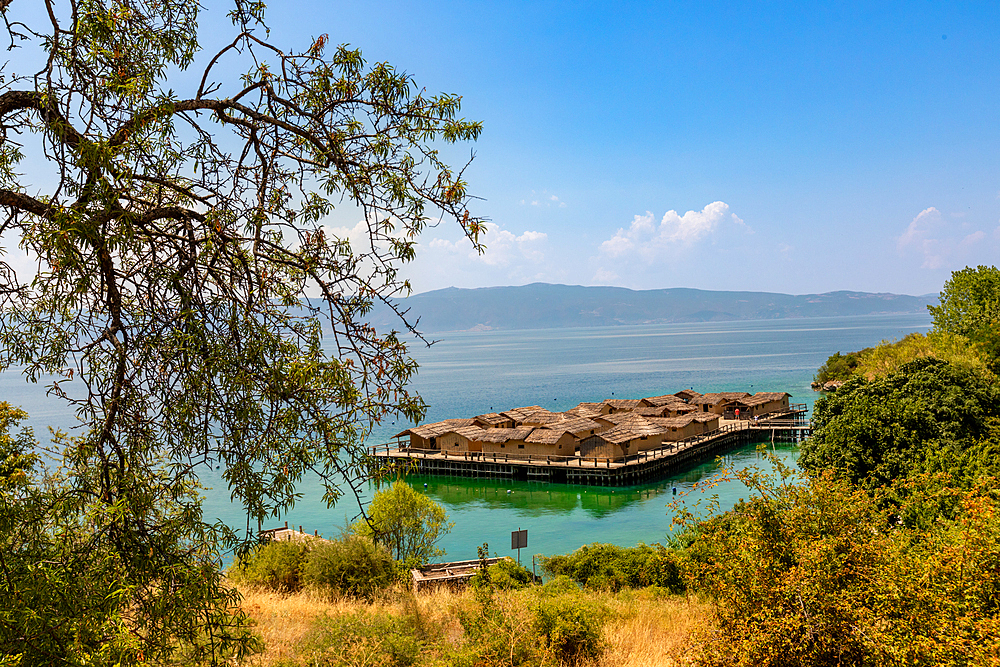Horizontal shot from an elevated viewpoint, partially framed by tree branches, showing a stilted village of thatched huts over turquoise water, with golden dry grass and shrubs in the foreground and distant mountains across the lake.
