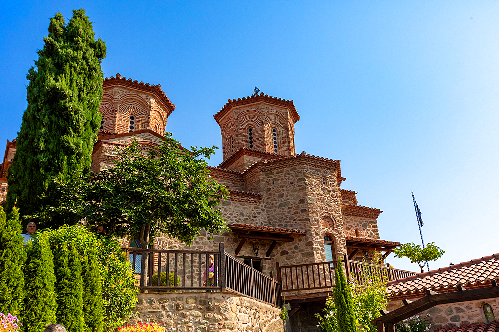 A historic stone monastery with twin Byzantine-style domes rises against a clear blue sky, surrounded by cypress trees, gardens, and wooden balconies.