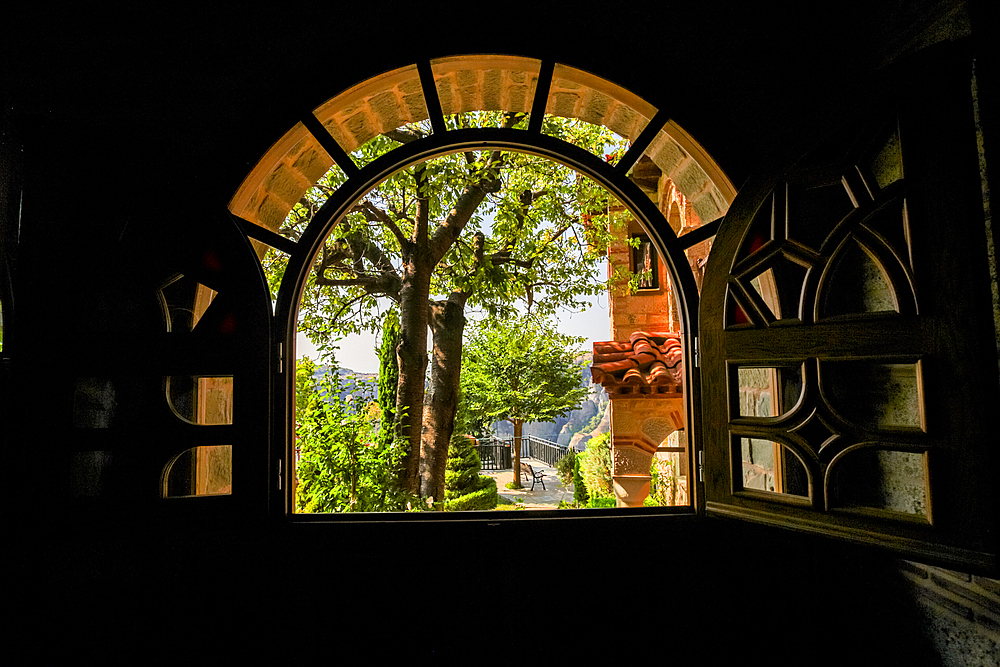 This image captures a scenic view framed by a large arched wooden window with open shutters. Through the window, you can see a sunlit courtyard with a tree, part of the monastery's tiled roof, and distant mountains under a bright sky.