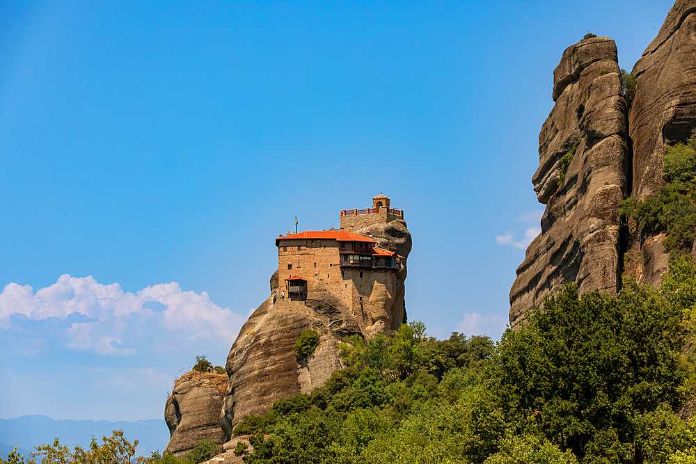 The Holy Monastery of Rousanou, dramatically situated on a narrow rock pillar, with its stone walls and red-tiled roof standing out against the rugged cliffs and bright blue summer sky in Meteora, Thessaly, Greece.