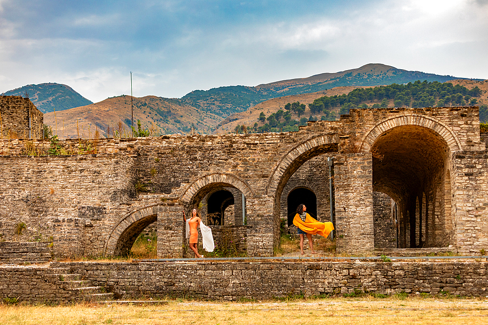Landscape orientation view of two women in colorful dresses posing among the stone archways of Gjirokastër Castle, with mountain scenery in the background, Gjirokastër, Albania.