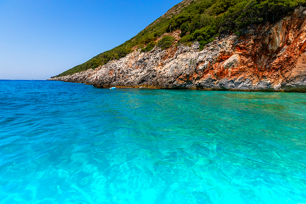 Clear turquoise waters of the Ionian Sea meeting rocky, vegetation-covered coastline near Dhermi, Vlorë County, Albania, on a bright summer day.