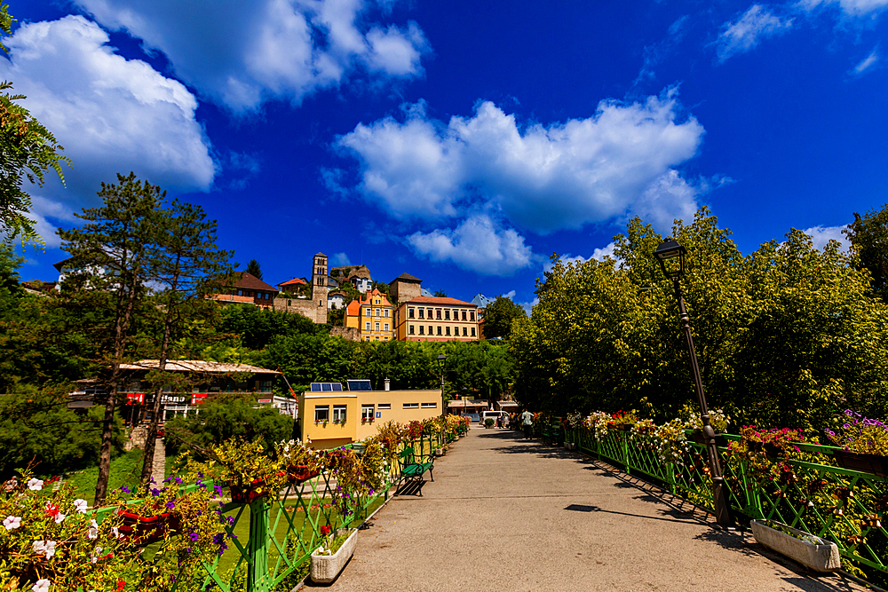 A flower-adorned pedestrian bridge in Jajce, Federation of Bosnia and Herzegovina, Bosnia and Herzegovina, leading to the old town with views of the Saint Luke bell tower and historic buildings under a bright summer sky.