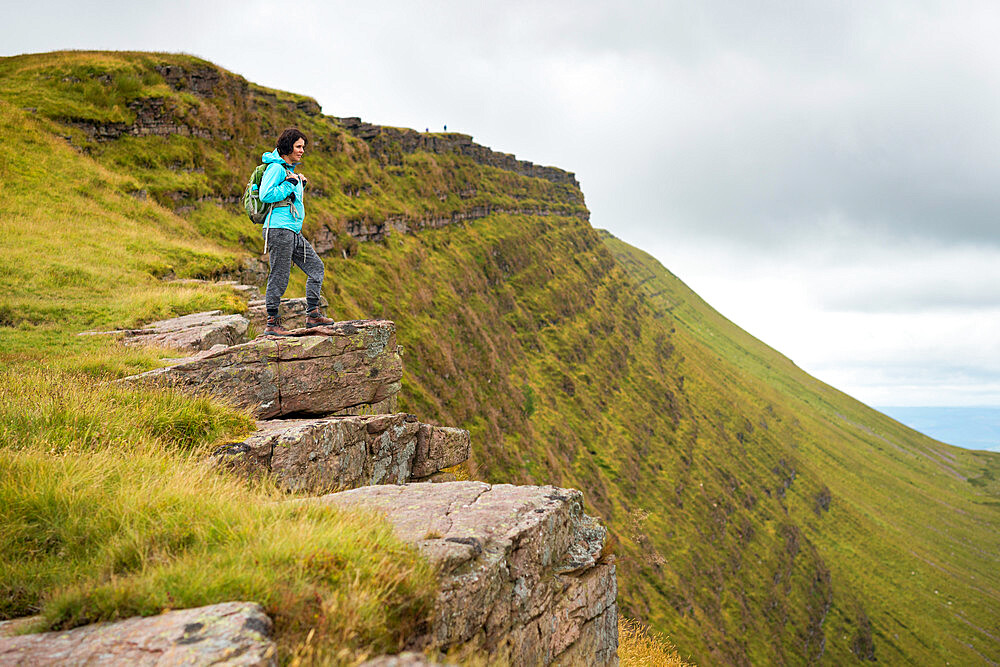 A woman looks out from a high escarpment while hiking in the Brecon Beacons National Park mountain range, Wales, United Kingdom, Europe