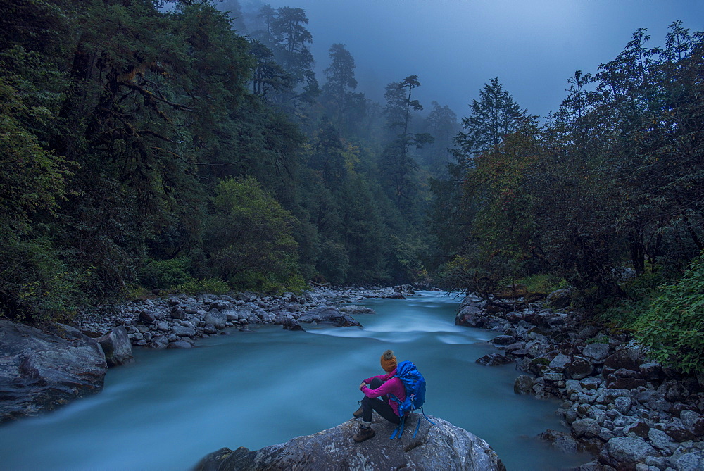 A woman takes a break from the trail and sits beside the Langtang Khola near the little village of Riverside on a misty evening, Nepal, Himalayas, Asia