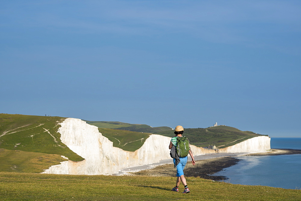 A woman walks along the chalk cliffs near Beachy Head with views of the Seven Sisters coastline in the distance, South Downs National Park, East Sussex, England, United Kingdom, Europe