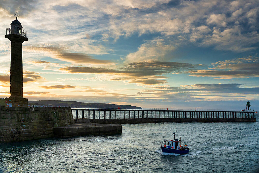 Sunset over Whitby harbour, pier and lighthouse as a small fishing boat comes into the harbour, Whitby, Yorkshire, England, United Kingdom, Europe