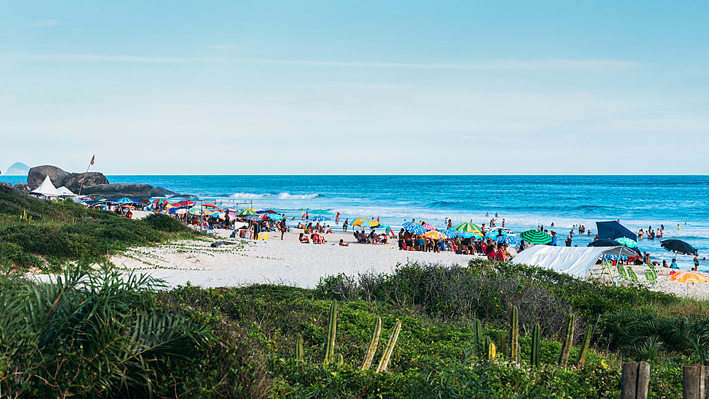 Crowds of beachgoers relax under colorful umbrellas on a sandy shoreline, taking in the ocean views and summer atmosphere, Grumari beach, Rio de Janeiro, Brazil, South America