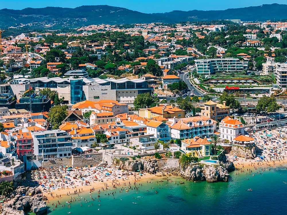 Aerial view of Cascais beaches and colourful rooftops of coastal town under bright sunlight with Sintra mountains in the background, Cascais, Portugal, Europe