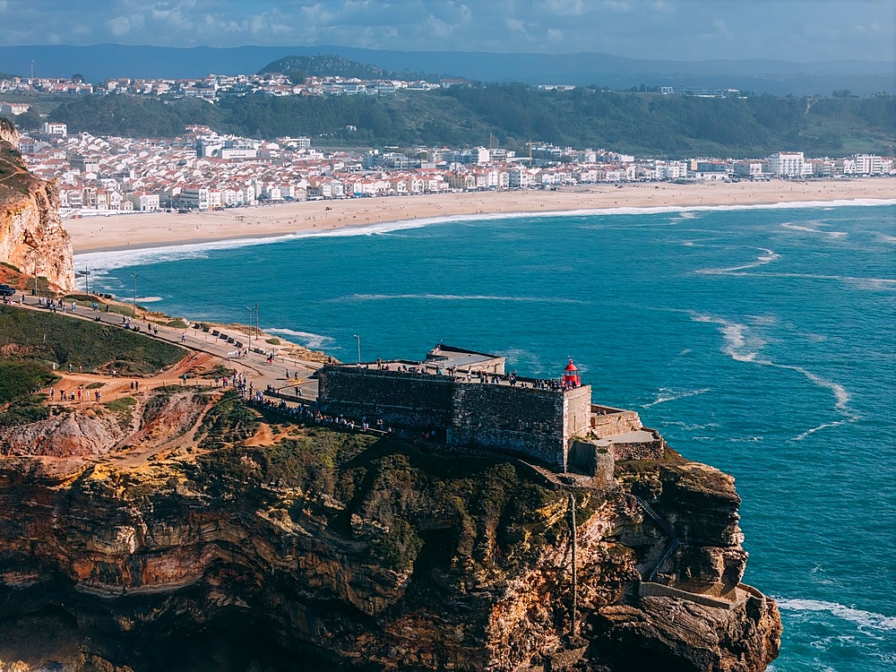 Tourists gather at the historic São Miguel Arcanjo lighthouse overlooking Nazaré's stunning coastline with giant waves, enjoying spectacular views of the ocean waves on a sunny day