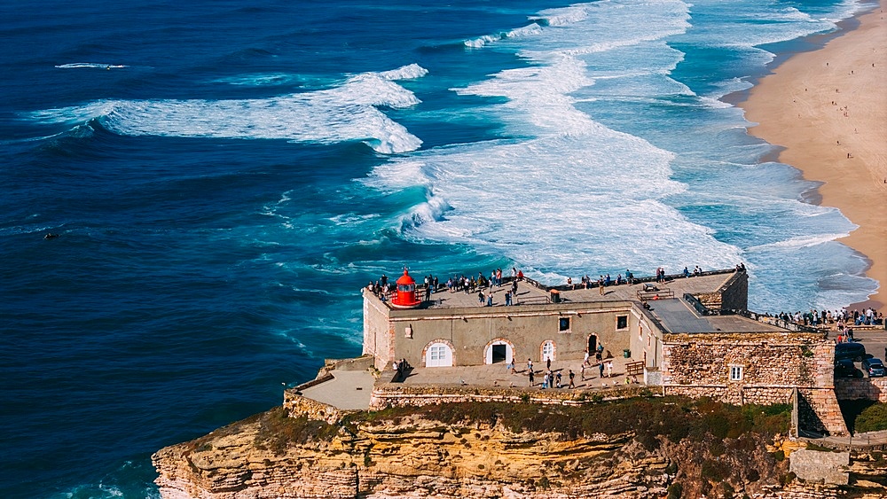 Tourists gather at the historic São Miguel Arcanjo lighthouse overlooking Nazaré's stunning coastline with giant waves, enjoying spectacular views of the ocean waves on a sunny day