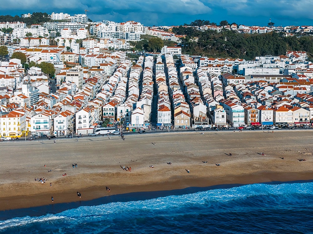 The lively beach of Nazaré, Portugal, revealing rows of charming houses with red roofs and people enjoying the sandy shore under a clear sky