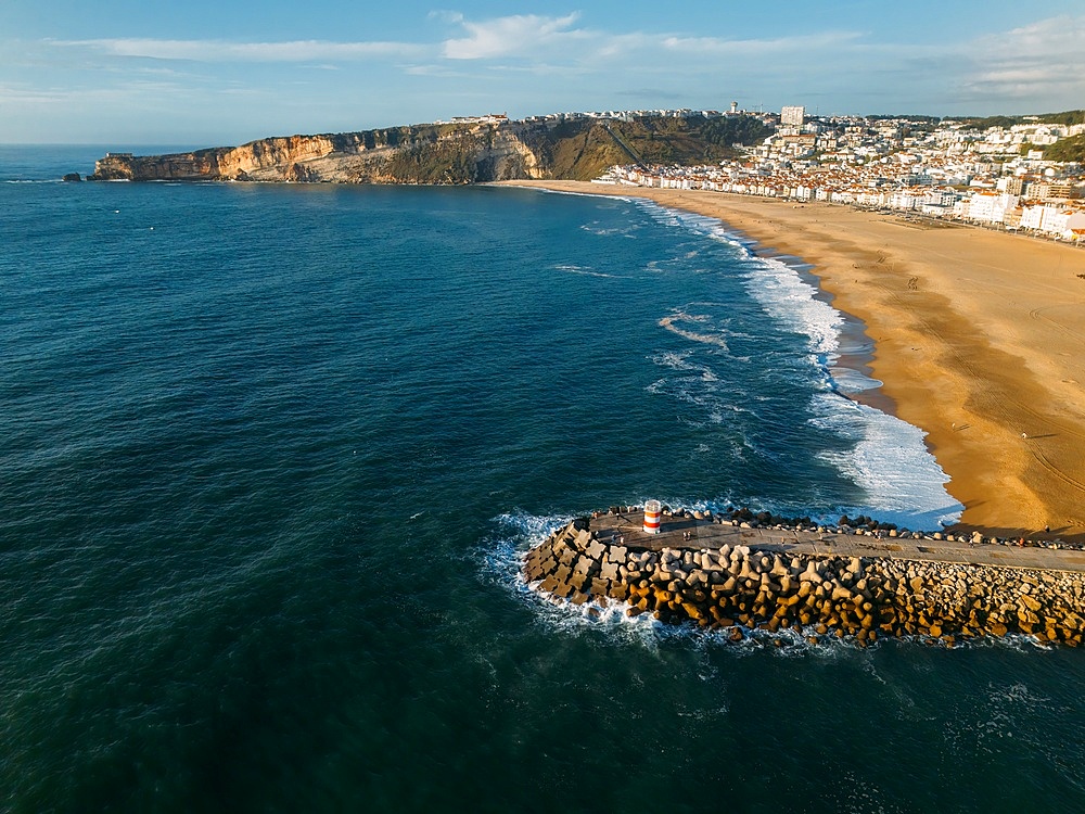 Aerial drone view of Nazare coastline showcasing beautiful beach, vibrant surf and rocky pier, Nazare, Oeste, Estremadura, Portugal, Europe