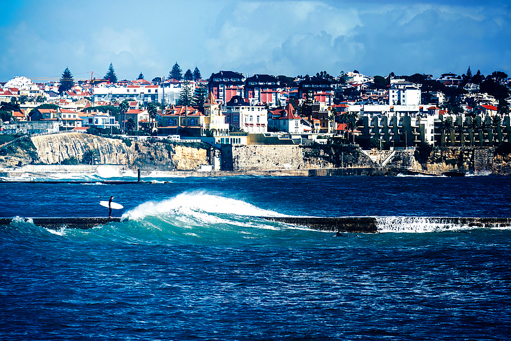 Surfer on a pier with stormy seas with Cascais/Estoril buildings in the background just outside of Lisbon, Portugal