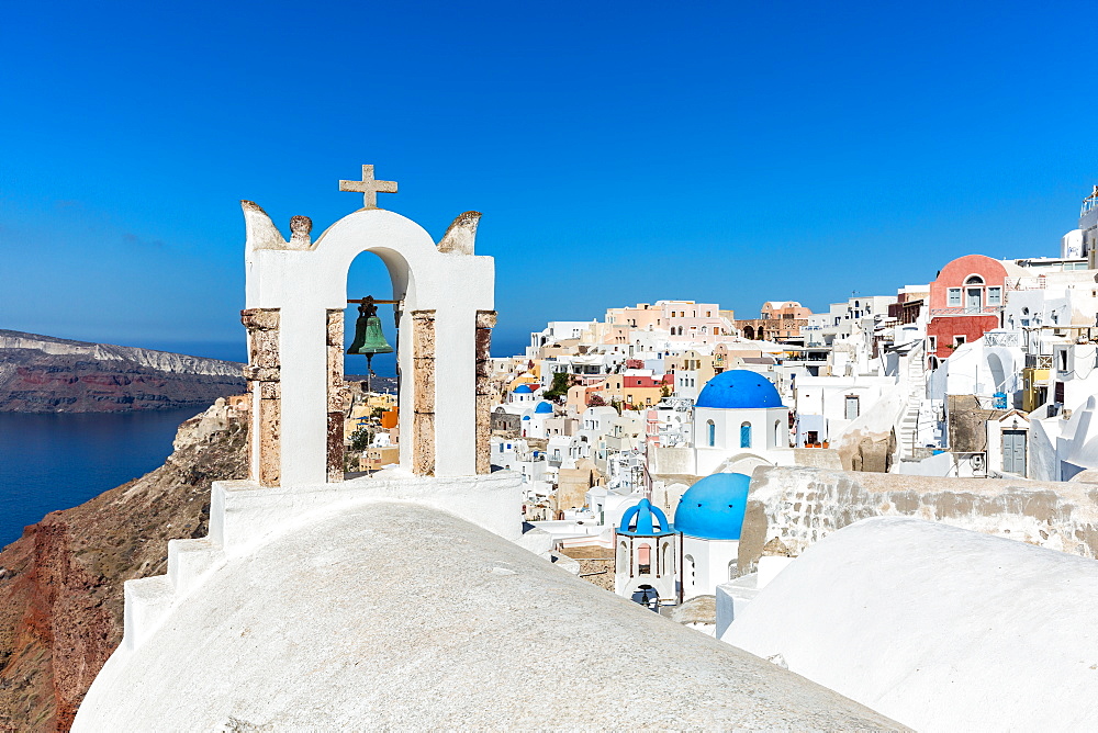 A church roof and bell with the white washed stone walls and blue church cupolas of Oia, Santorini, Cyclades, Greek Islands, Greece, Europe