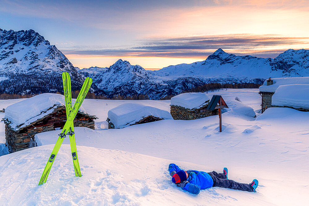 Young skier relaxes in the snow with view of the small village at sunrise, Valmalenco, Valtellina, Lombardy, Italy, Europe