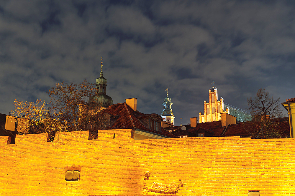 Night low angle view of illuminated defensive wall fortification in front of traditional low-rise roof-tiled houses and passing clouds above in the Old Town of Warsaw, Poland, Europe