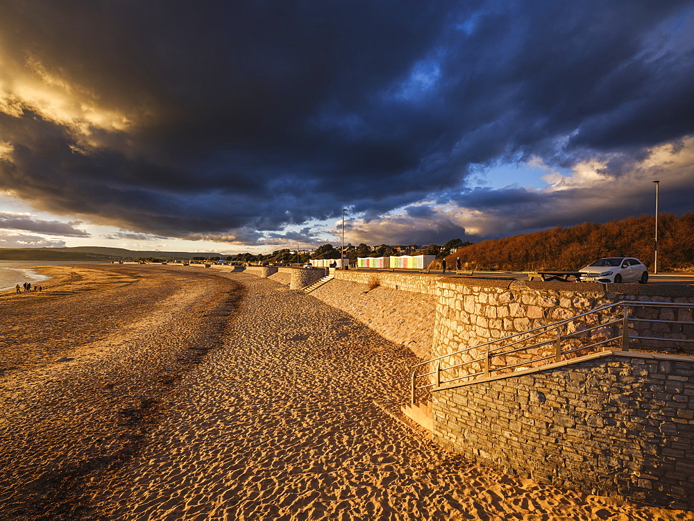 Under dramatic clouds, strong warm sunlight illuminates the beach and sea defences on the sea front at Exmouth, Devon, England, United Kingdom, Europe