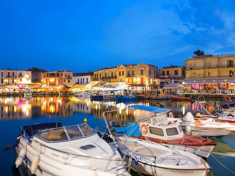 View across the Venetian Harbour at dusk, brightly lit restaurants reflected in water, Rethymno (Rethymnon), Crete, Greek Islands, Greece, Europe