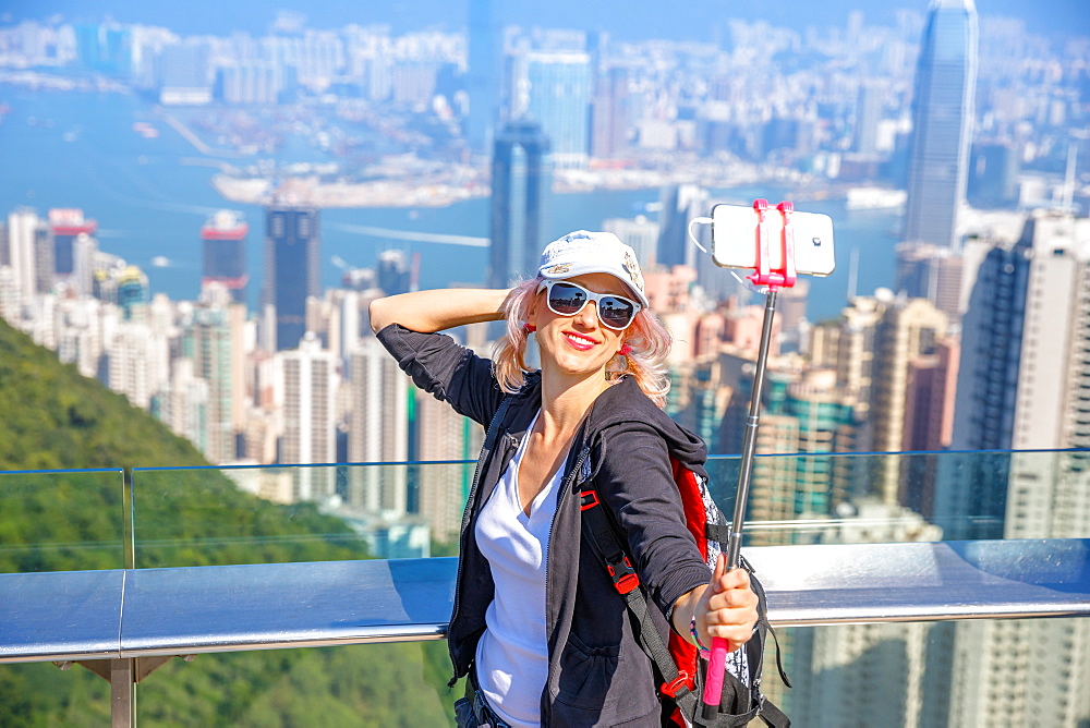 Tourist taking selfie stick picture photo with smart phone enjoying view over Victoria Harbour from viewing platform on top of Peak Tower, Victoria Peak, Hong Kong, China, Asia