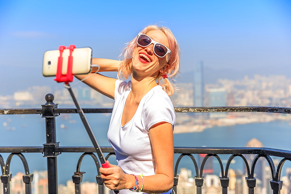 Tourist taking selfie stick picture with smart phone, enjoying view over Victoria Harbour from viewing platform on top of Peak Tower, Victoria Peak, Hong Kong, China, Asia