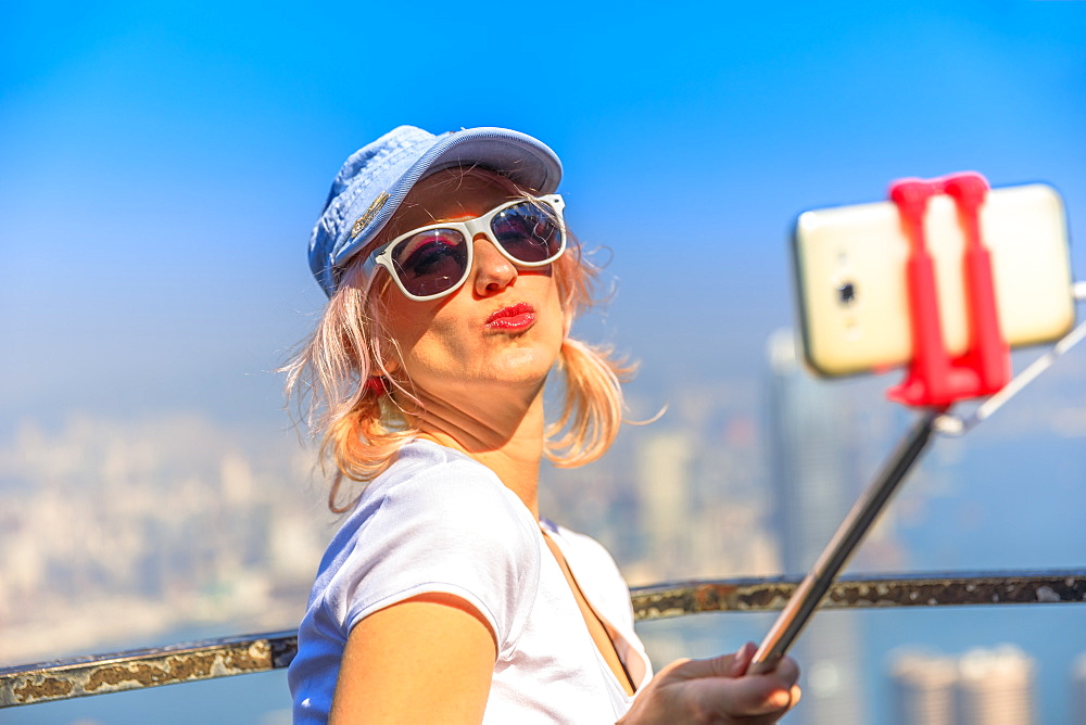 Tourist taking selfie stick picture with smart phone, enjoying view over Victoria Harbour from viewing platform on top of Peak Tower, Victoria Peak, Hong Kong, China, Asia