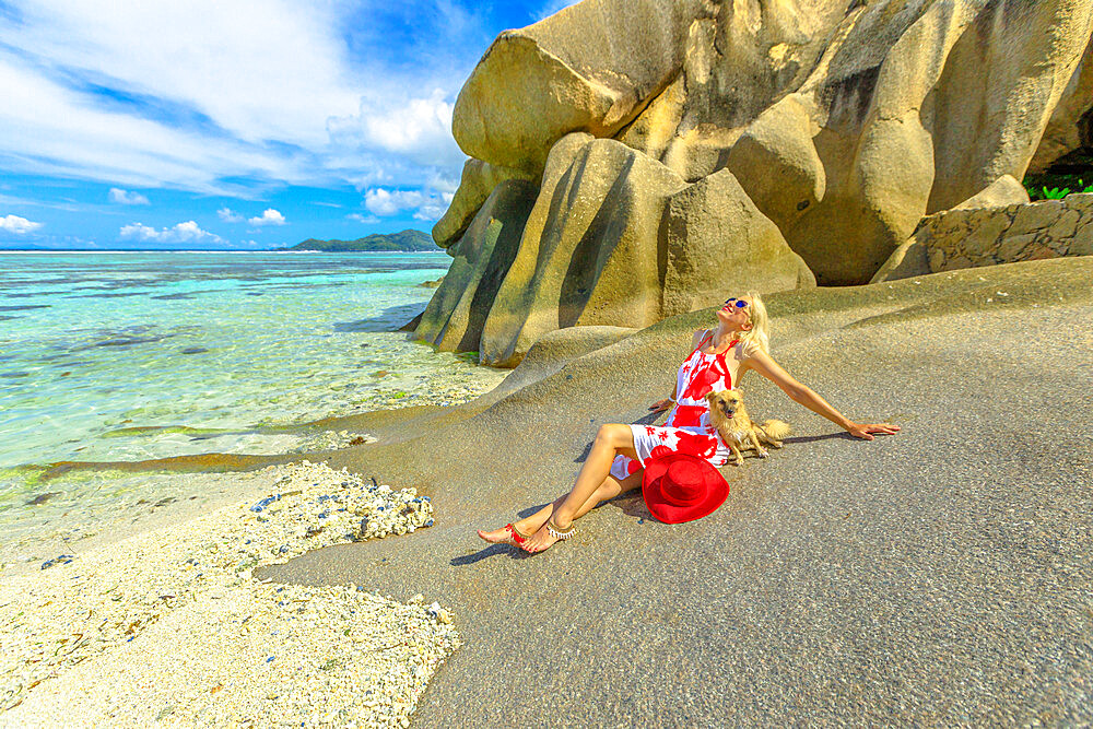 Tourist woman and cute dog sitting on beach at low tide, with granite rocks behind, at Anse Source d'Argent, La Digue, Seychelles, Indian Ocean, Africa