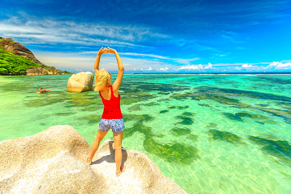 Tourist woman with raised arms in red dress standing on a huge boulder at Anse Source d'Argent, La Digue, Seychelles, Indian Ocean, Africa