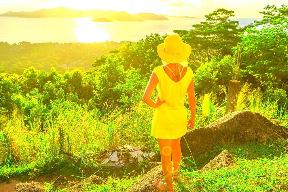 Tourist woman in yellow dress enjoying view of Praslin Island from top viewpoint of La Digue at sunset, Seychelles, Indian Ocean, Africa