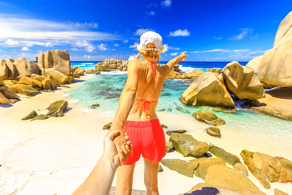 Blonde tourist woman in orange bikini holding hand of partner at Anse Marron, La Digue island, Seychelles, Indian Ocean, Africa