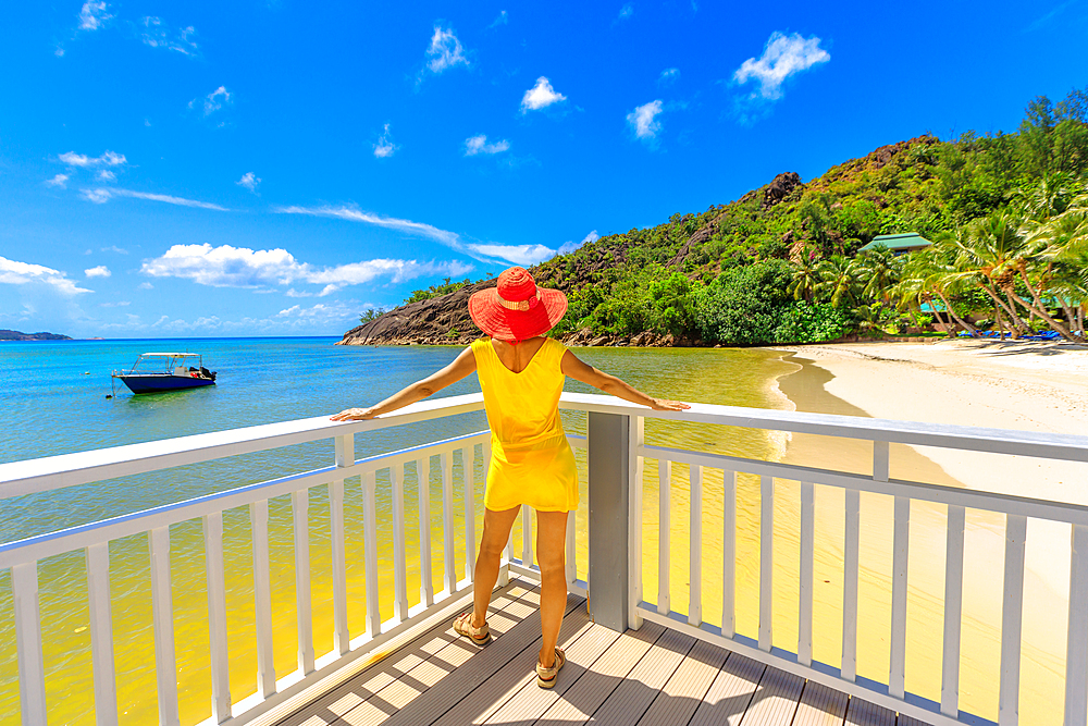 Woman in yellow dress on balcony of wooden jetty, looking at pristine white beach of Anse Gouvernement, near Cote d'Or Bay, in Praslin, Seychelles, Indian Ocean, Africa