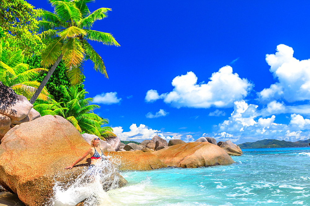 Tourist woman in splashing wave at Felicite Island, boulders and palm trees behind, La Digue, Seychelles, Indian Ocean, Africa