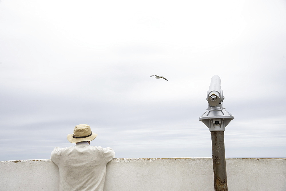 Man peering over a wall beside a telescope along the Algarve coast of southern Portugal, Europe