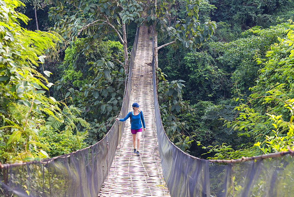 A woman crosses a precarious looking suspension bridge over the jungle in Laos, Indochina, Southeast Asia, Asia