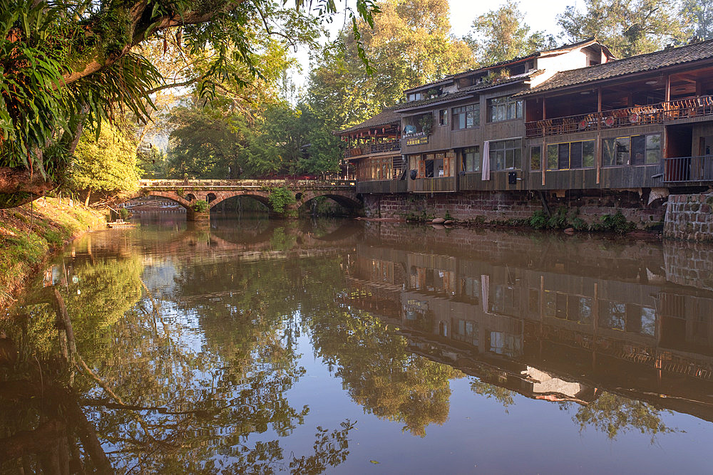A three-arched stone bridge surrounded by vegetation in Shangli Town at dawn and its reflection in the river.