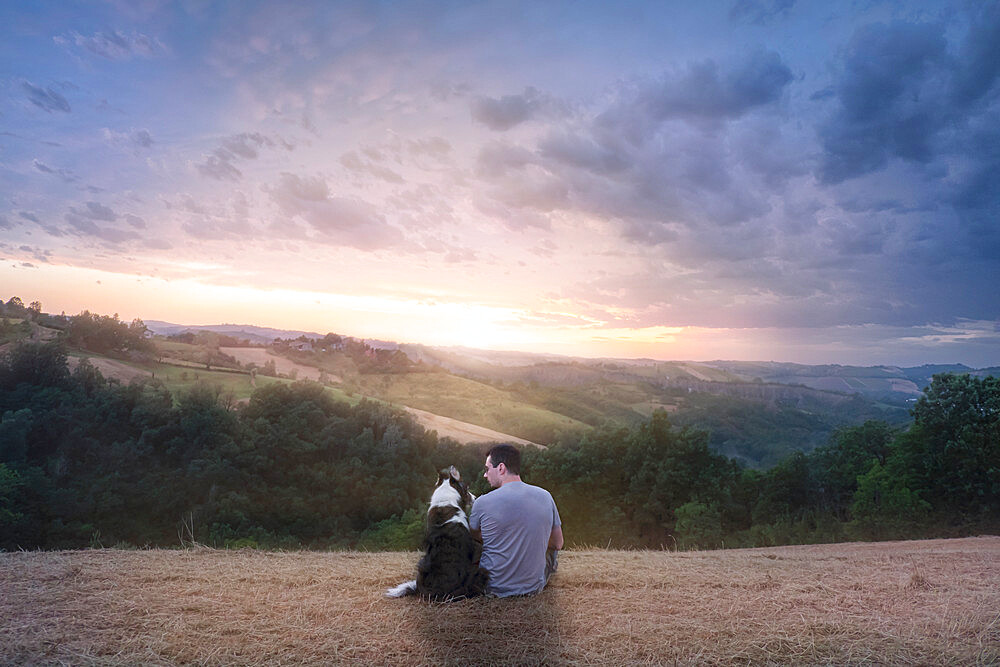 A man and his border collie dog sitting in a field in the countryside enjoying sunset, Italy, Europe