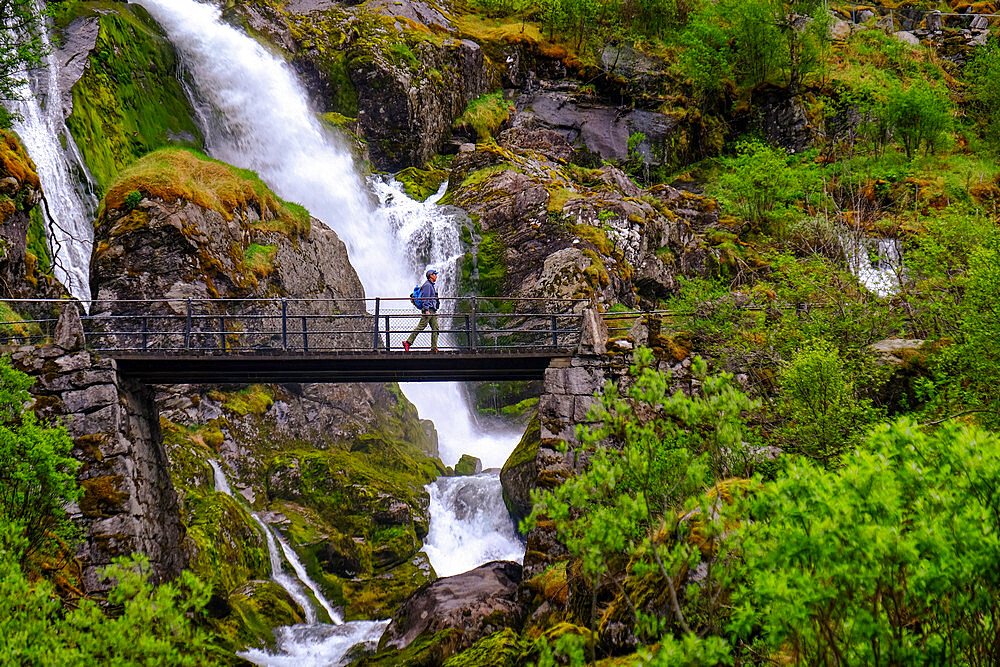 A man on a pedestrian bridge crosses one of many waterfalls originating at the Briksdal glacier, Stryn, Vestland, Norway, Scandinavia, Europe