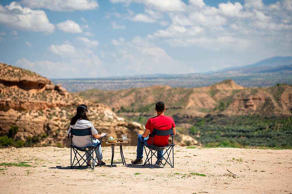 Couple sit on chairs with a table looking at a desert like landscape near Francisco Abellan Dam, Granada, Andalusia, Spain, Europe