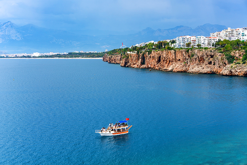 Antalya city view near the cliffs with a tourist boat on the sea with turquoise water and mountains in the background, in Turkey