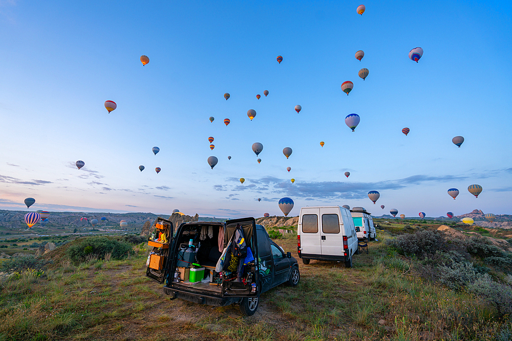 Camper vans parked in Love valley in front of hot air balloons at sunrise in Cappadocia, Turkey