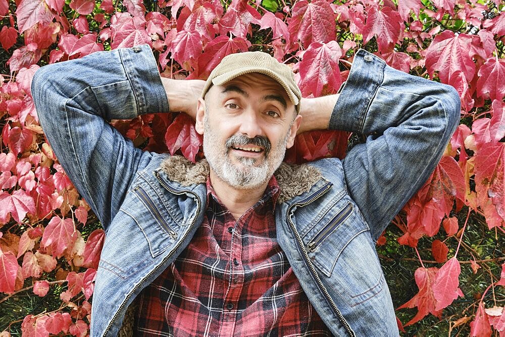 Portrait of a mature caucasian man dressed in a checked shirt, denim jacket and a cap with an autumnal leaves background.