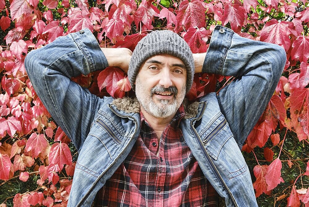 Portrait of a mature caucasian man dressed in a checked shirt, denim jacket and a wool cap with an autumnal leaves background.