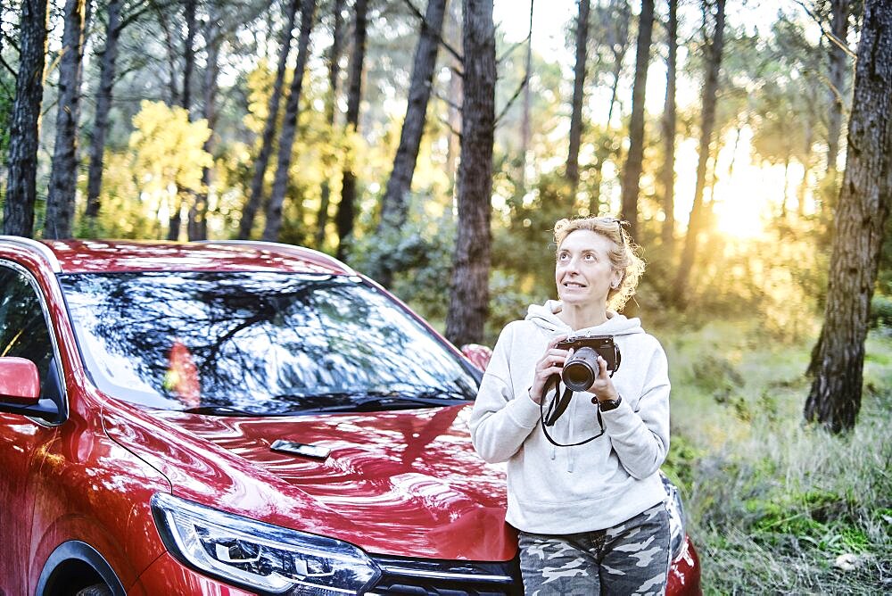 Young adult caucasian woman taking pictures at sunset close to a car parked in a forestry path in autumn. Travels concept. Ayegui, Navarre, Spain, Europe.