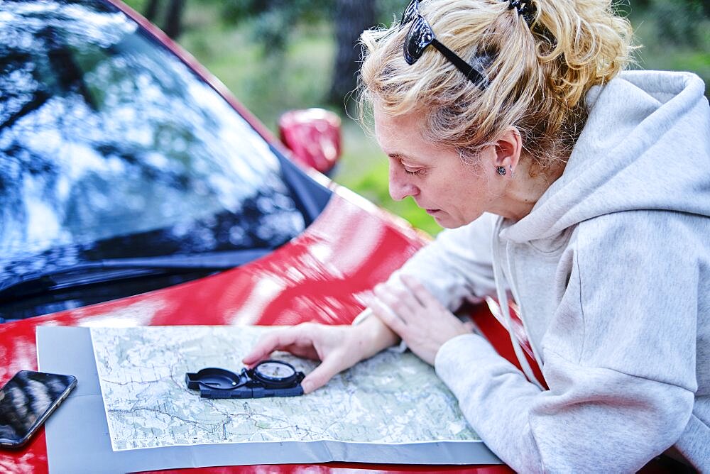 Young adult caucasian woman orienting herself with a map and a compass close to a car parked in a forestry path. Travels concept. Ayegui, Navarre, Spain, Europe.