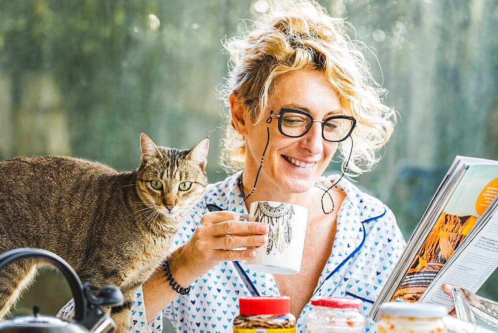 Blonde young mature woman with glasses in pijamas at home in breakfast time, reading a magazine and having a moment with her cat.