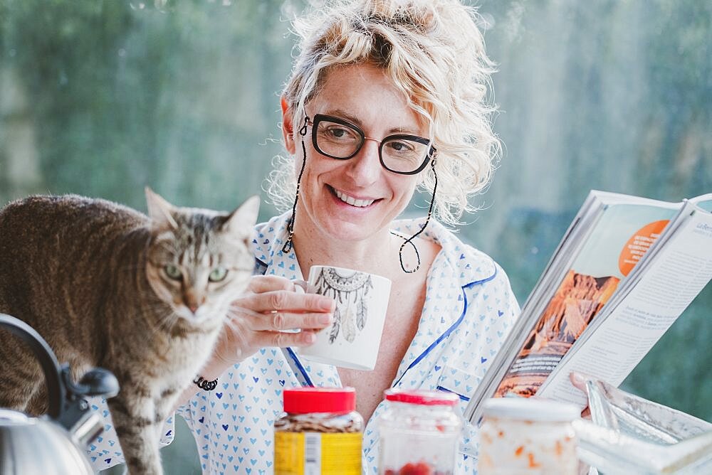 Blonde young mature woman with glasses in pijamas at home in breakfast time, reading a magazine and having a moment with her cat.