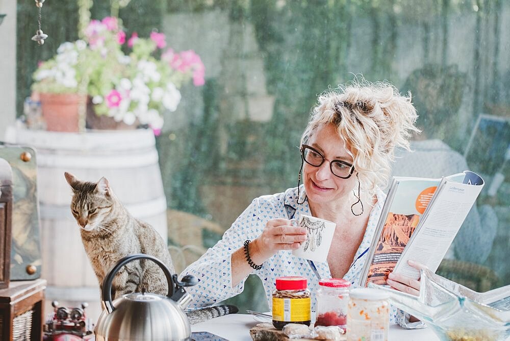 Blonde young mature woman with glasses in pijamas at home in breakfast time, reading a magazine and having a moment with her cat.