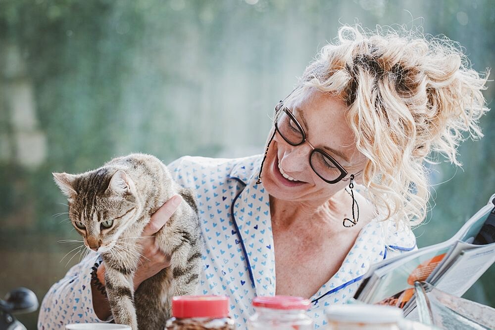 Blonde young mature woman with glasses in pijamas at home in breakfast time, reading a magazine and having a moment with her cat.